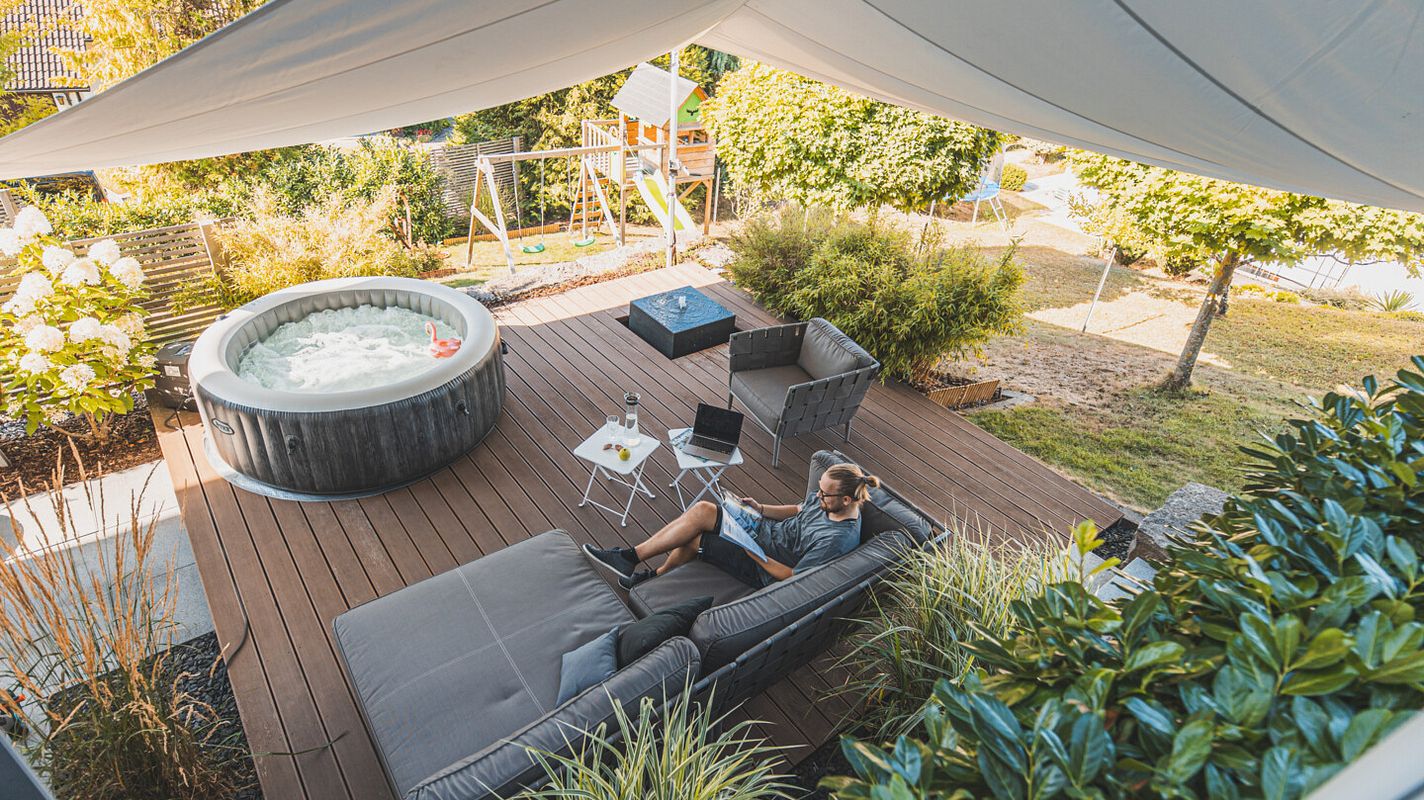 Eine gemütlich eingerichtete Holzterrasse mit Möbeln und Whirlpool unter einem großen Sonnensegel